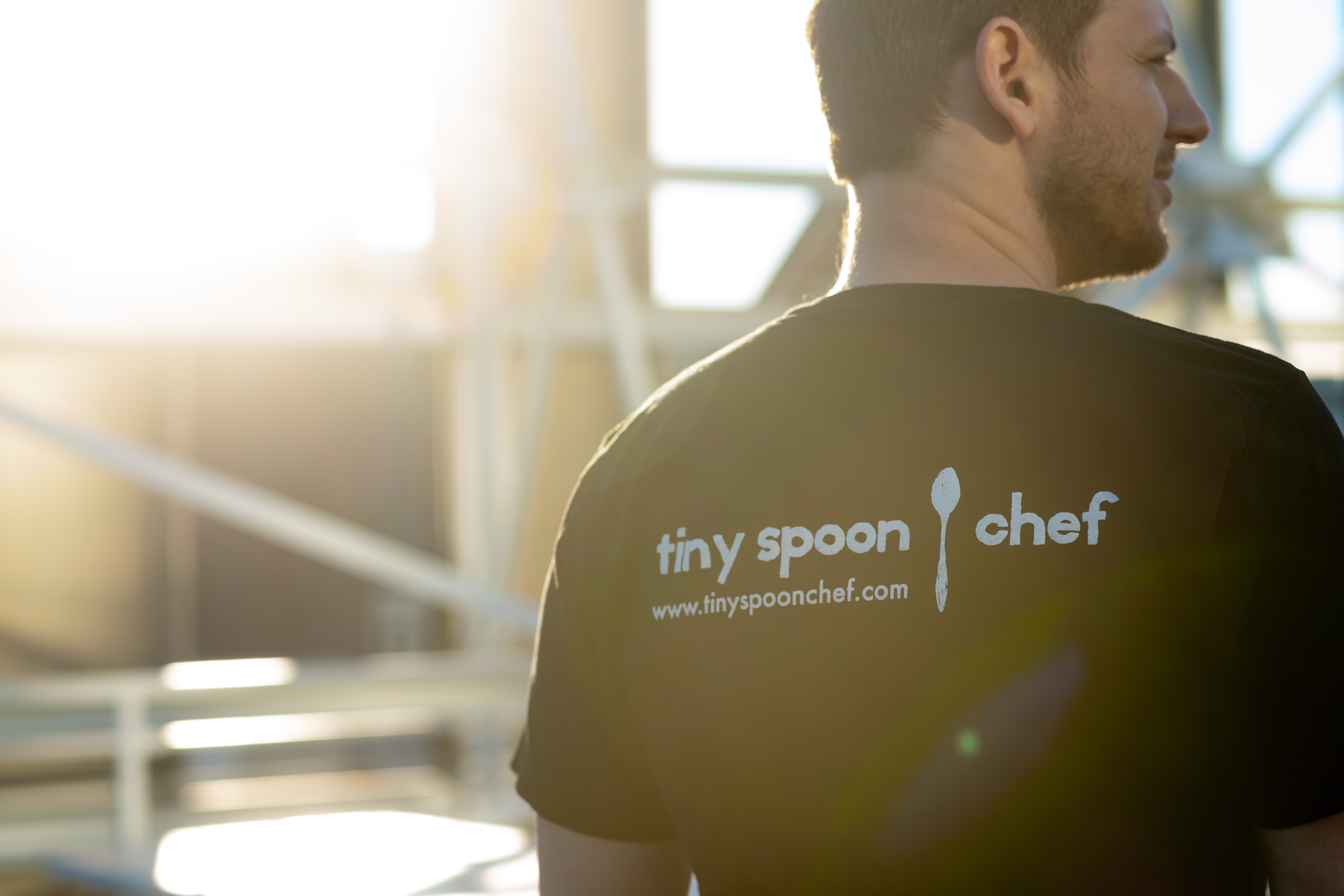 Tiny Spoon chef in a branded T-shirt, seen from behind in warm sunlight